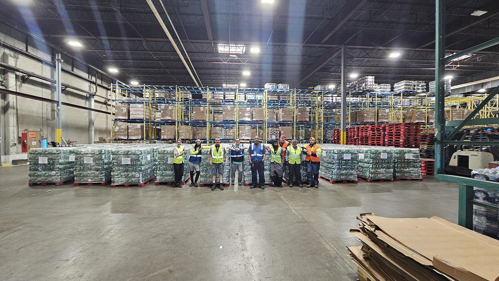 A diverse group of individuals stands in a warehouse surrounded by stacked boxes, engaged in conversation and planning durring Hurricane Helene.